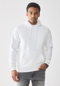 Man met een effen witte hoodie met een voorzak en trekkoorden, grijze jeans en een zwart horloge, die tegen een witte achtergrond staat.