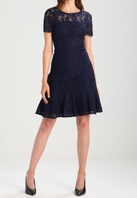 Abito in pizzo blu navy con maniche corte, vita aderente e orlo svasato indossato con tacchi neri a punta su sfondo uniforme.