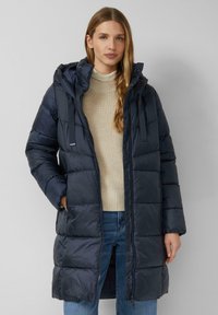 Doudoune marine avec capuche, coutures verticales et fermeture éclair. Portée sur un pull en maille beige clair, associée à un jean bleu.