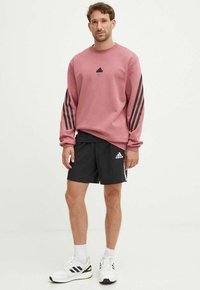 Felpa rosa con design a tre righe nere sulle maniche e logo nero sul petto; abbinata a pantaloni corti neri con accenti bianchi e logo.