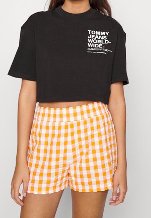 Camiseta negra recortada con texto blanco "Tommy Jeans World Wide" combinada con pantalones cortos de talle alto a cuadros naranjas y blancos en una persona de pie.