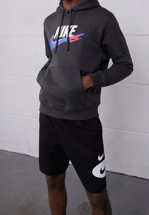 Personne portant un sweat à capuche noir Nike avec des swooshes bleus et rouges, un short noir Nike avec des swooshes blancs, debout les mains dans la poche du sweat.