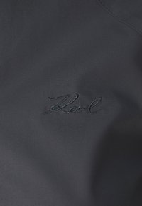 Σκούρο ναυτικό ύφασμα με κεντημένη υπογραφή "Karl" σε χρώμα αντίστοιχο του υφάσματος, παρουσιάζοντας λεία υφή και διακριτικό σχέδιο.