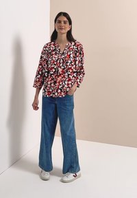 Femme debout, une main dans la poche, portant un chemisier floral rouge et rose, un jean large bleu et des baskets blanches dans un studio simple.