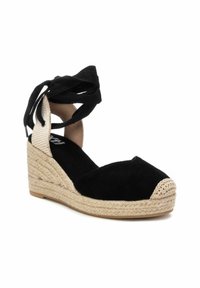 Zapato de cuña de ante negro con suela de cuerda trenzada y punta redondeada. Presenta tiras de tela negra entrelazadas alrededor del tobillo.