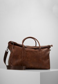 Braune Leder-Duffel-Tasche mit Reißverschluss, doppelten Griffen, verstellbarem Schultergurt und sichtbaren Texturmuster auf der Oberfläche.