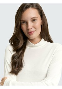 Jeune femme aux longs cheveux bruns portant un pull à col roulé blanc, bras croisés, regardant l'appareil photo avec un sourire doux.