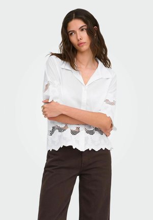 Vrouw met golvend donker haar, draagt een witte blouse met kanten rand en korte mouwen en een donkere broek, staat met de armen over elkaar en kijkt opzij.