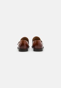 Braune Lederloafers mit glänzendem Finish, niedrigem Profil und kleinem Absatz, mit einem subtilen Slipper-Design und dekorativen Hardware-Details.