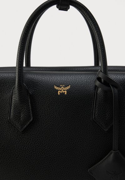 Μαύρη υφασμάτινη δερμάτινη τσάντα χειρός με στρογγυλεμένες λαβές, χρυσό λογότυπο MCM και δερμάτινη ετικέτα που κρέμεται από λουράκι στη δεξιά πλευρά.