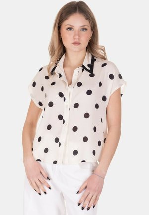 Donna in piedi che indossa una camicetta color crema a maniche corte con pois neri e pantaloni bianchi, in posa con le mani vicino ai fianchi.