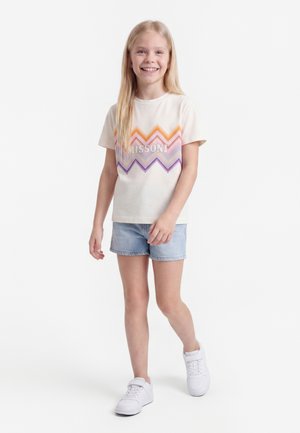 Χαμογελαστή νεαρή κοπέλα με μακριά ξανθά μαλλιά, που φοράει ένα λευκό μπλουζάκι Missoni με ζιγκ-ζαγκ σχέδιο, ανοιχτόχρωμα τζιν σορτς και λευκά αθλητικά παπούτσια.