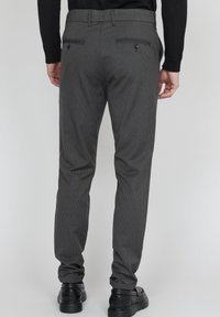 Man som bär grå randiga kostymbyxor och svarta skor, står med ryggen mot en enkel ljus bakgrund.