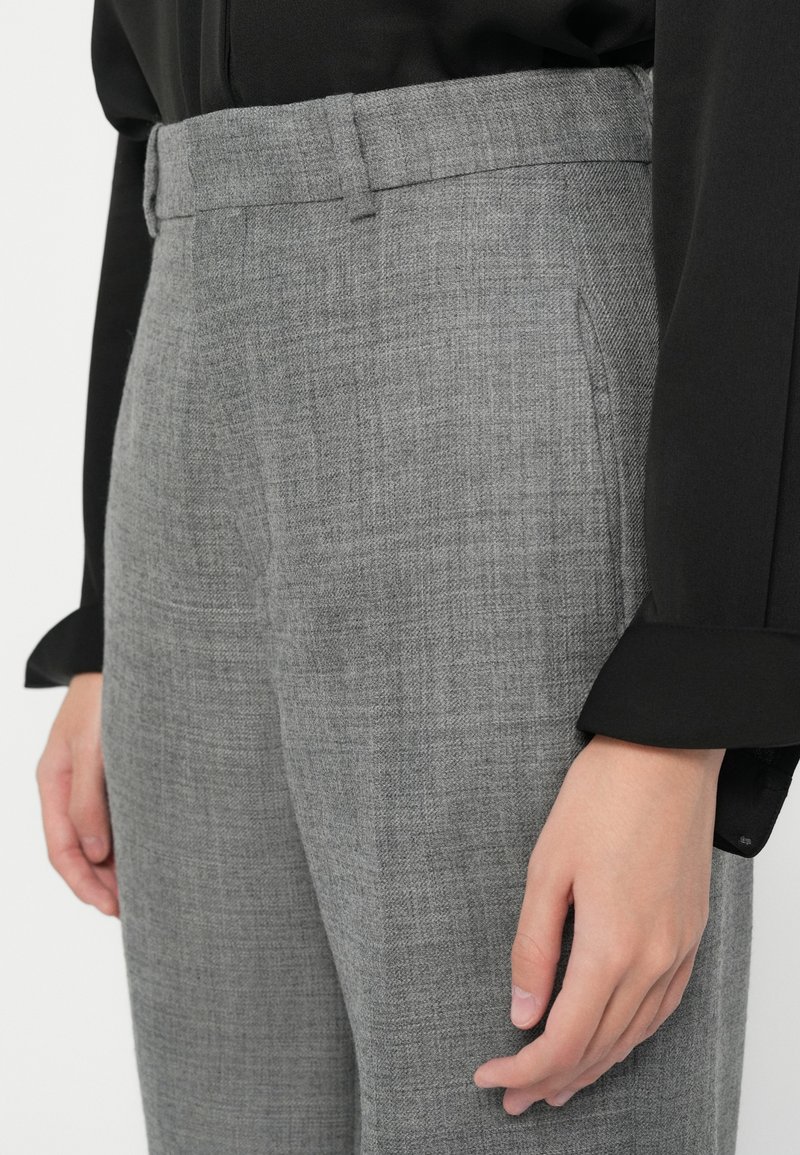 Primo piano di una persona che indossa pantaloni grigi tessuti e una maglietta nera a maniche lunghe, con le mani rilassate ai lati.