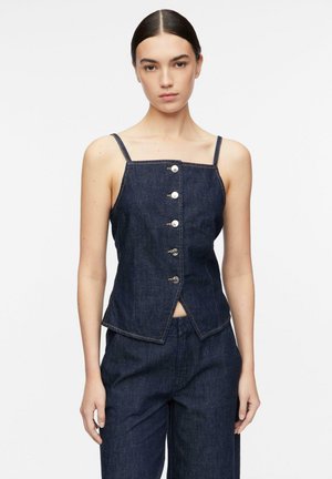 Kobieta ubrana w ciemnoniebieski, dżinsowy, bezrękawnikowy top na guziki z cienkimi ramiączkami i pasujące spodnie z wysokim stanem na białym tle.