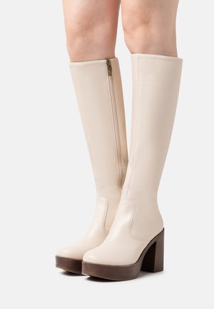 Bottes crème en similicuir montant jusqu'au genou, avec une fermeture éclair latérale, un bout arrondi et un talon bloc en bois de 5 pouces pour plus de hauteur.