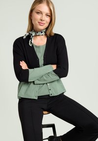 Cardigan nero sopra una maglia verde, con scollatura a barca. La modella indossa una sciarpa a fantasia. Pantaloni neri e uno sgabello di legno completano l'outfit.