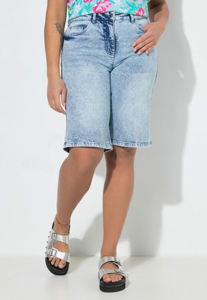 Person trägt hellblaue, knieumspielende Jeansshorts, ein farbenfrohes, florales Oberteil und silberne Sandalen mit Schnalle, steht vor einem weißen Hintergrund.