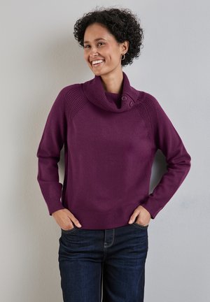 Violetti neulepaita, jossa on kauluksen omaava kaulus ja sivupainikkeet, resorireunukset hihoissa ja helmassa. Yhdistetty tummiin farkkuihin, neutraalilla taustalla.