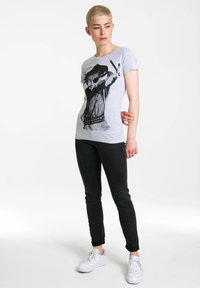 Grå bomuld t-shirt med korte ærmer, der har et sort grafisk motiv af en person, der laver et fredstegn. Bæres med sorte jeans og hvide sneakers.
