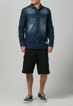 Camisa de mezclilla en azul oscuro con parte frontal desteñida, diseño de botones y dos bolsillos en el pecho, combinada con pantalones cortos cargo negros y zapatillas grises.