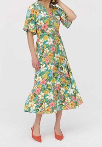 Robe maxi florale à manches courtes, avec un fond turquoise et des fleurs vibrantes jaunes, roses et blanches ; taille cintrée et ourlet évasé.