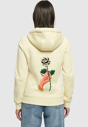 Keltainen huppari, jossa on käden graafinen kuvasto pitämässä valkoista ruusua, jossa on vihreät lehdet. Pehmeä kangas ja rento istuvuus hupulla.