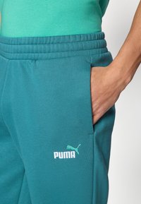 Παντελόνια φόρμας σε τυρκουάζ χρώμα με κορδόνι στη μέση, πλαϊνή τσέπη και μικρό λευκό λογότυπο Puma. Μαλακή, λεία υφή, κατάλληλη για καθημερινή χρήση.