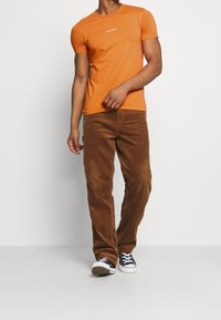 Oranžové bavlněné tričko s krátkými rukávy a malým bílým logem na hrudi, spárované s hnědými kordovými kalhotami a černými teniskami.