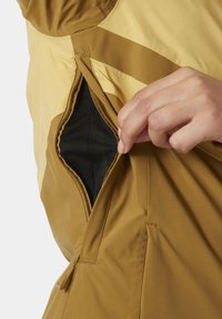 Wasserabweisende Jacke in Gelb und Braun, mit einer Reißverschlusstasche mit schwarzem Netzeinsatz. Glatte Textur und dynamische Designelemente.