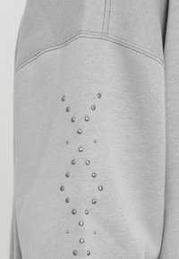Felpa grigia con un tessuto testurizzato, dotata di un pannello liscio e una linea decorativa di borchie metalliche lungo la manica.