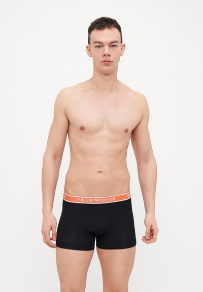 Boxers noirs avec une ceinture orange Emporio Armani, présentant une coupe ajustée et un tissu lisse, présentés sur un modèle avec un fond neutre.