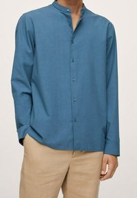 Camicia a maniche lunghe con bottoni di colore blu medio, dotata di colletto alla cinese, tessuto morbido e una leggera lucentezza, abbinata a pantaloni beige chiaro.
