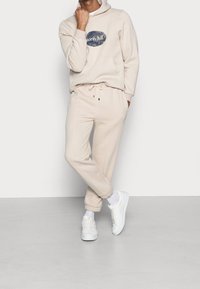 Beige hoodie with a front pocket and navy logo, paired with matching joggers. White sneakers and crew socks complete the outfit.