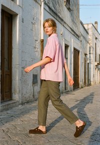 Jeune femme aux cheveux blonds marche dans une rue pavée en pierre, portant une chemise rose, un pantalon olive et des chaussures marron sans lacets, à la lumière vive du jour.