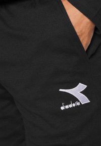 Mustat puuvillaiset collegehousut, joissa on valkoinen brodeerattu logo vasemmassa taskussa, yksinkertainen muotoilu ja pehmeä texture.