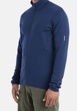 Mann trägt eine enganliegende marineblaue Jacke mit seitlichen Reißverschlusstaschen und olivgrüne Hose vor schlichtem Hintergrund.