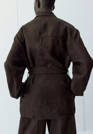 Persona con chaqueta marrón oscuro con cinturón y pantalones a juego, de pie con las manos en las caderas frente a una pared blanca lisa.