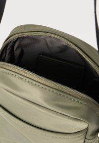 Πράσινη τσάντα ώμου από νάιλον με φερμουάρ, που ανοίγει αποκαλύπτοντας εσωτερικό από πιο σκούρο ύφασμα και ορατή μαύρη θήκη καρτών στο εσωτερικό.