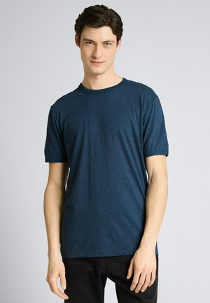 Jeune homme aux cheveux foncés portant un t-shirt à manches courtes bleu foncé uni et un pantalon noir, debout devant un fond clair uni.