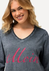 Maglietta a maniche lunghe grigia con scollo a V, caratterizzata da una texture liscia e da audaci lettere in corsivo rosa che formano la scritta "Moin". Design semplice con sottili variazioni di colore.