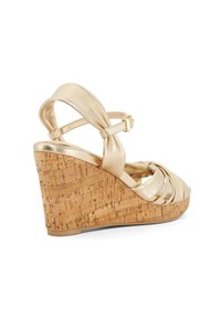 Gouden metallic wedge sandaal met kurken plateau. Beschikt over kruislings gespen en een enkelgesp. Gladde textuur en taps toelopend ontwerp verbeteren de stabiliteit.