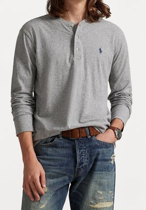 Homme portant un t-shirt henley gris à manches longues avec un logo bleu, une ceinture en cuir marron, un jean bleu déchiré et une montre-bracelet avec un bracelet en cuir marron.