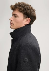 Cappotto di lana grigio scuro con colletto alto, chiusura con un singolo bottone e tessuto testurizzato. Presenta una toppa rotonda nera con logo sulla manica.