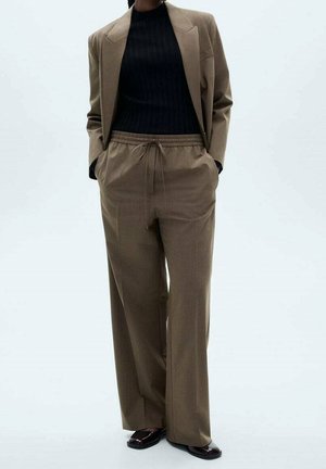 Personne portant un blazer marron et un pantalon à cordon assorti avec un col roulé côtelé noir et des mocassins foncés, debout les mains dans les poches.