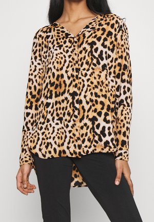 Kobieta ubrana w bluzkę z nadrukiem w panterkę z długim rękawem i kołnierzykiem, połączoną z czarnymi spodniami, stojąca na jednolitym tle.