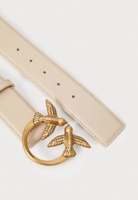 Beige leren riem met een ronde goudkleurige gesp, ontworpen met twee gedetailleerde vogels. De riem heeft zichtbare gaatjes voor aanpassing.
