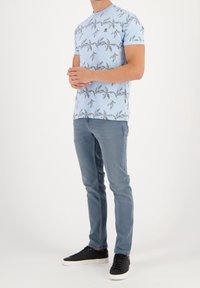 Muž oblečený v modrém tričku s květinovým vzorem, modrých slim-fit džínách a černých teniskách s bílou podrážkou, stojí s sepjatýma rukama.