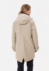 Beige waterdichte jas met capuchon, ontworpen met een strakke silhouet, verstelbare manchetten en een langere achterzoom voor extra dekking.