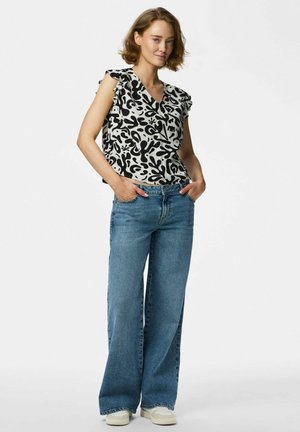 Donna con capelli corti castani che indossa una maglia con maniche a volant a motivo bianco e nero, jeans a gamba larga blu e sneakers bianche, con le mani nelle tasche.